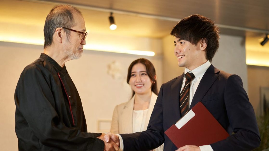 握手をするスーツを着た男性二人とそれを見ている女性