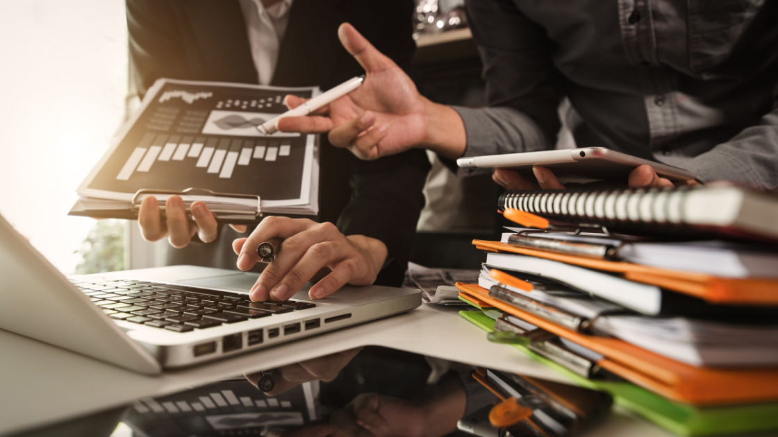 ノートパソコンを操作するビジネスマンと、机の上に積み上げられた紙の資料