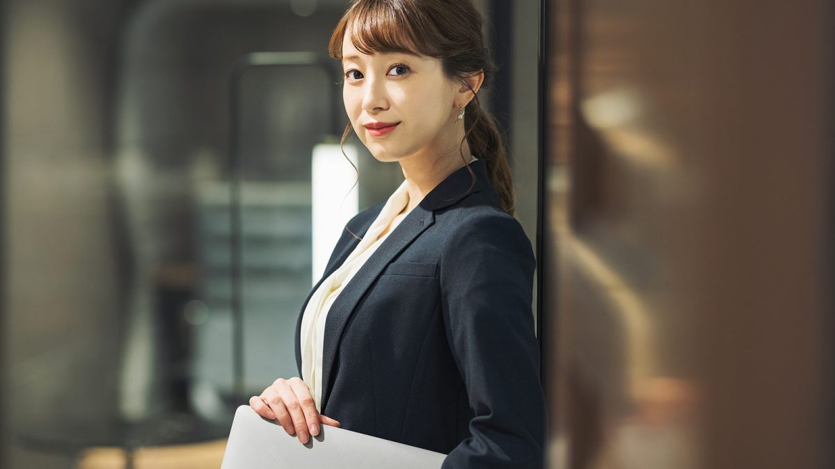 脇にパソコンを抱えて立つスーツ姿の女性