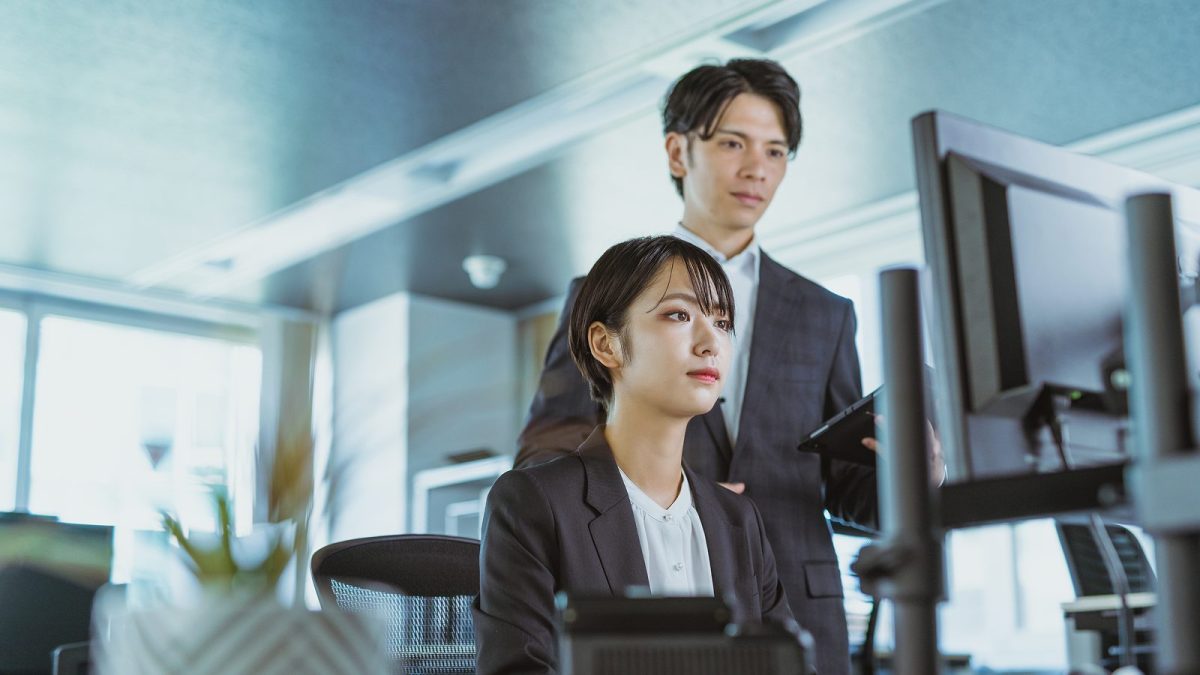 座ってパソコンを操作する女性と立っている男性