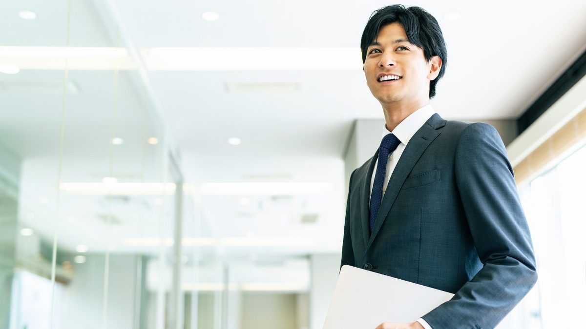 ノートパソコンを小脇に抱えてオフィスの廊下を歩く男性