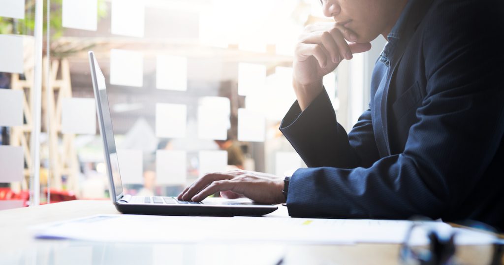 ノートパソコンを操作しながら顎に手を当てるビジネスマン