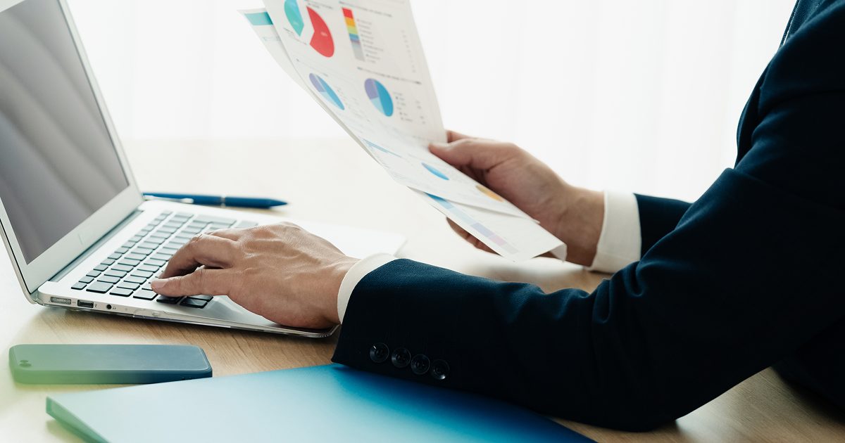 紙の資料を片手に持ち、ノートパソコンを操作する様子