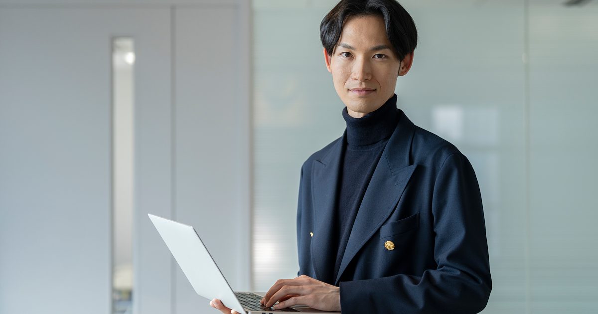 ノートパソコンを片手に持ち、こちらを見る男性