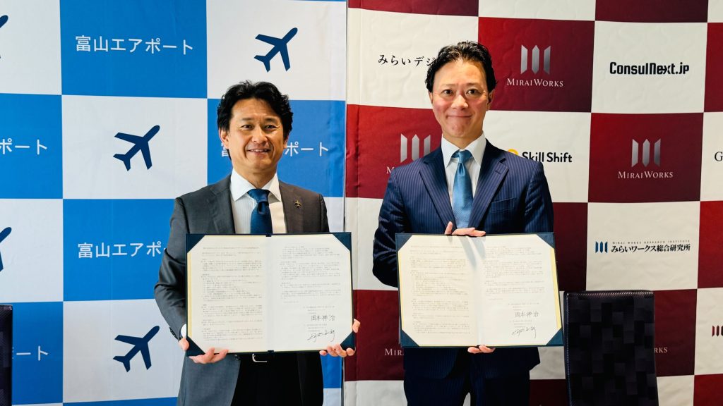 富山エアポートのロゴ背景の前に立つ岡田信一郎社長（左）と、みらいワークスのロゴ背景の前に立つ岡本祥治社長（右）。締結された連携協定書を両手で広げて持ち、笑顔で正面を向いている写真。